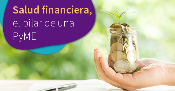 Mano sosteniedo un envase de vidrio lleno de monedas y coronado con una planta verde, salud financiera, mano sobre escritorio, hojas y pluma, fondo verde desenfocado
